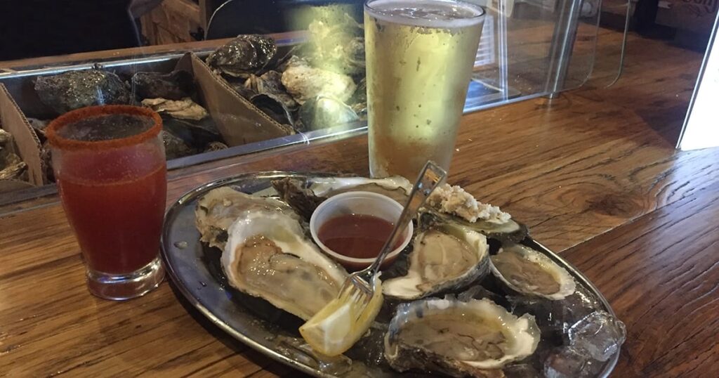 A metal tray of raw oysters with lemon and sauce sits on a wooden bar next to a glass of beer and a chili-rimmed red cocktail—just one highlight at the best restaurant in Woodbine MD, known for top-notch service and catering in Woodbine MD.