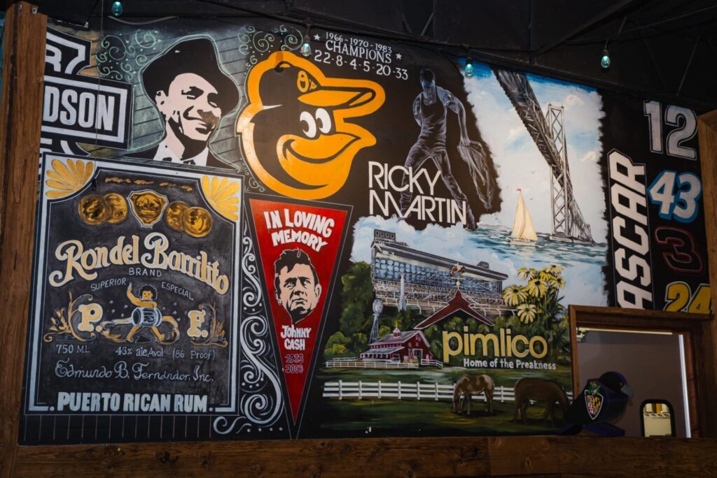 A colorful mural features portraits, a rum ad, sports themes, a sailboat, the Pimlico racetrack, and an In Loving Memory tribute—celebrating culture and history as vibrantly as Latin American food in Woodbine MD.