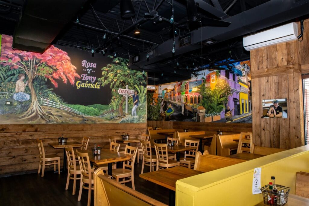 A colorful restaurant interior with wooden tables and chairs, murals of vibrant trees and lively street scenes, and condiment bottles on tables. Enjoy delicious Latin American food in Woodbine MD in this empty, well-lit space.