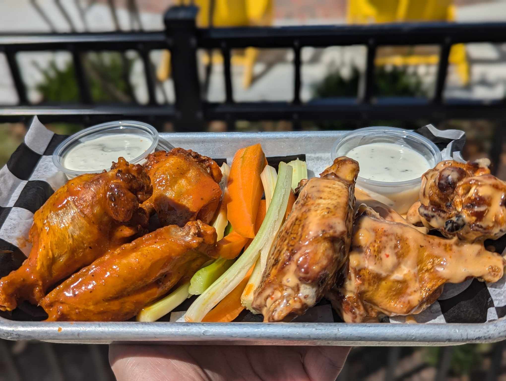 A tray of buffalo wings and sauced chicken wings with ranch, plus carrot and celery sticks, is served outdoors—perfect for enjoying classic American food in Woodbine MD. A black fence and yellow decor blur in the background.