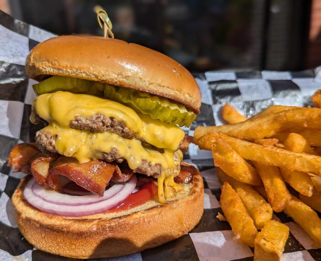 Enjoy classic American food in Woodbine MD with our cheeseburger—two beef patties, melted cheese, bacon, pickles, red onion, and tomato on a toasted bun, served with crispy French fries on checkered paper.