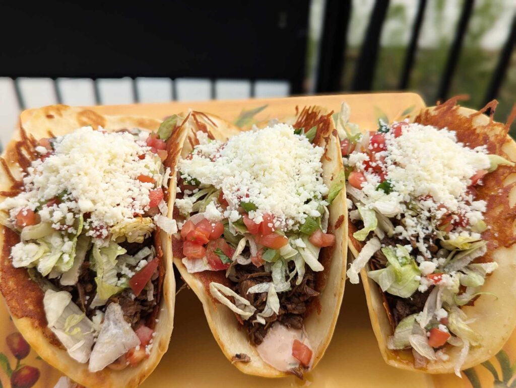 Three crispy tacos filled with shredded beef, lettuce, diced tomatoes, and crumbled cheese are arranged on a plate—perfect for anyone craving authentic Latin American food in Woodbine MD. A blurred outdoor background completes the inviting scene.